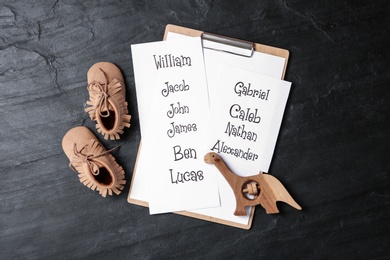 Clipboard with list of baby names, child's shoes and wooden toy on black table, flat lay