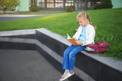 Photo of Cute little girl with backpack reading textbook on city street. Space for text