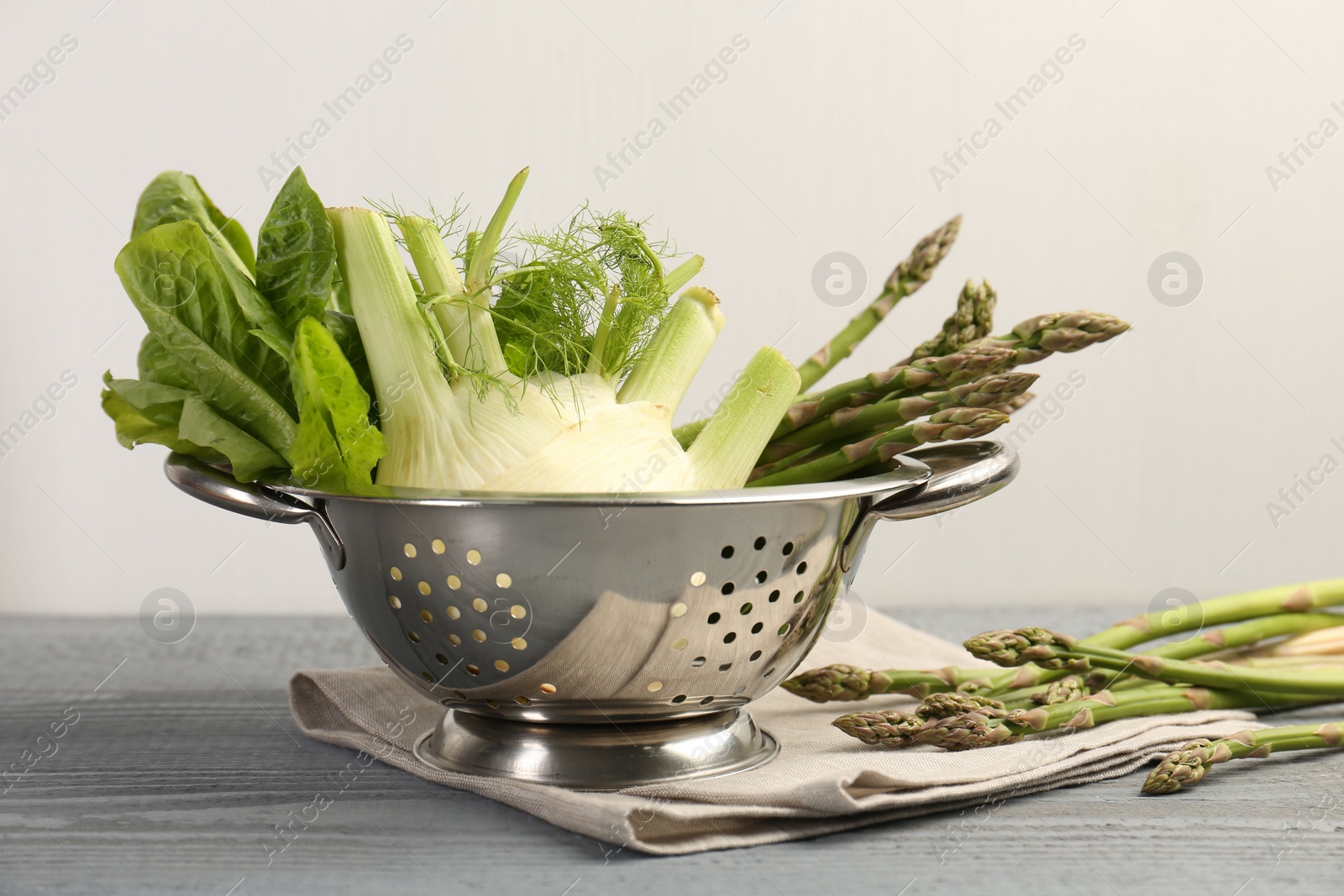 Photo of Metal colander with fennel, lettuce and asparagus on gray wooden table