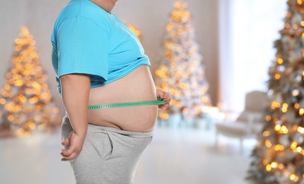 Image of Overweight man measuring his waist in room with Christmas trees after holidays, closeup