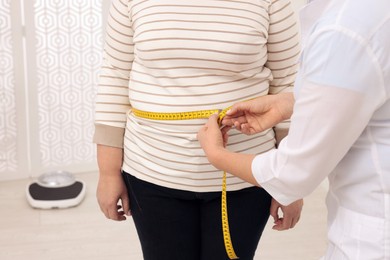 Photo of Nutritionist measuring overweight woman's waist with tape in clinic, closeup