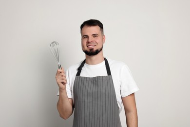 Happy professional confectioner in apron holding whisk on light grey background