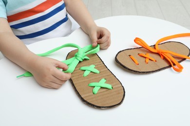 Photo of Little boy tying shoe lace using training cardboard template at white table, closeup