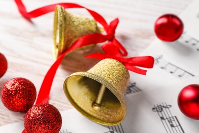 Photo of Golden shiny bells with red bows, music sheets and decorative balls on wooden table, closeup. Christmas decoration