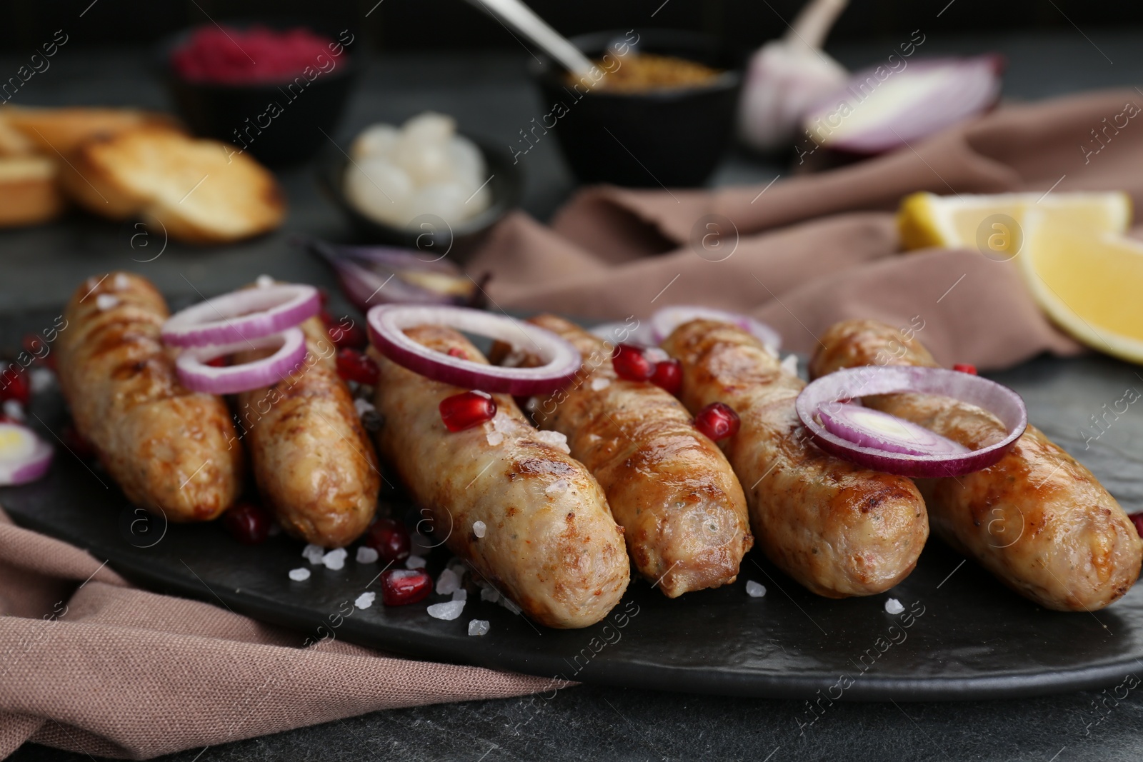 Photo of Tasty grilled sausages with onion rings and pomegranate seeds served on grey table, closeup