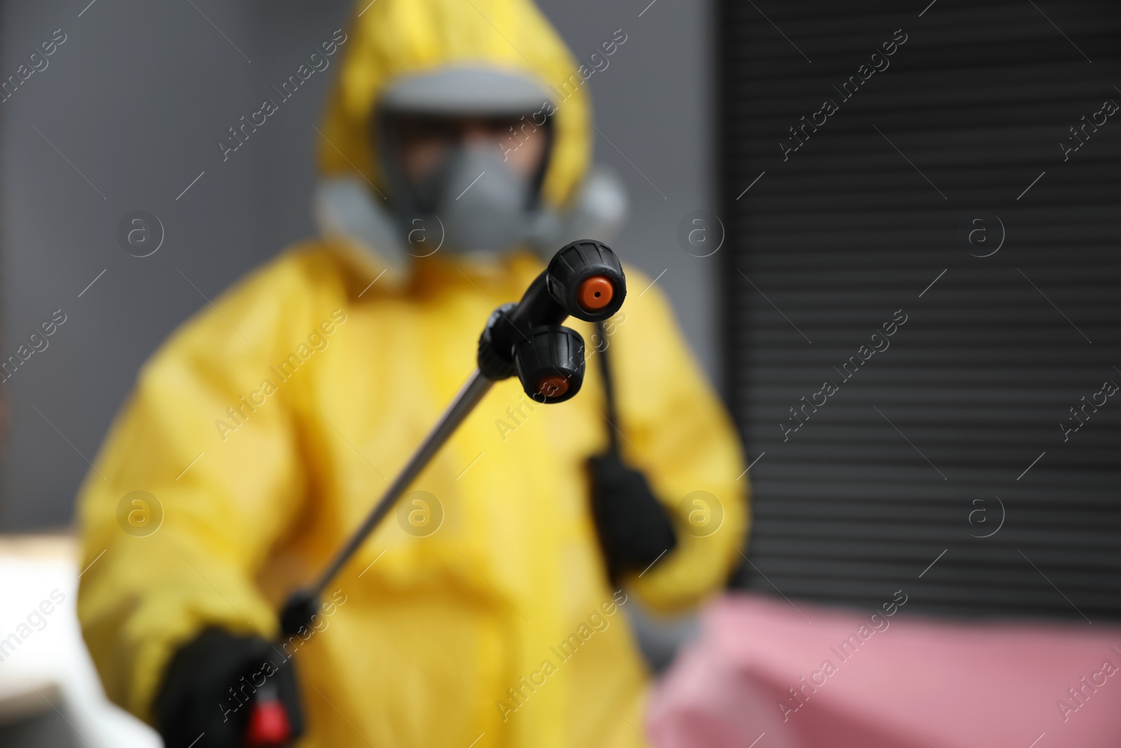 Photo of Pest control worker in protective suit indoors, focus on insecticide sprayer