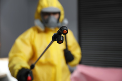 Photo of Pest control worker in protective suit indoors, focus on insecticide sprayer