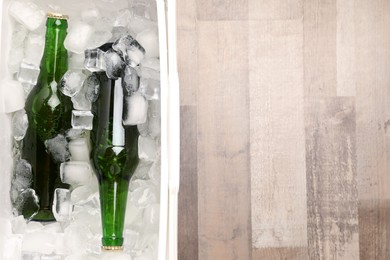 Plastic cool box with ice cubes and beer on wooden floor, top view. Space for text