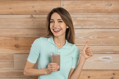 Young woman with glass of chocolate milk showing thumb up on wooden background