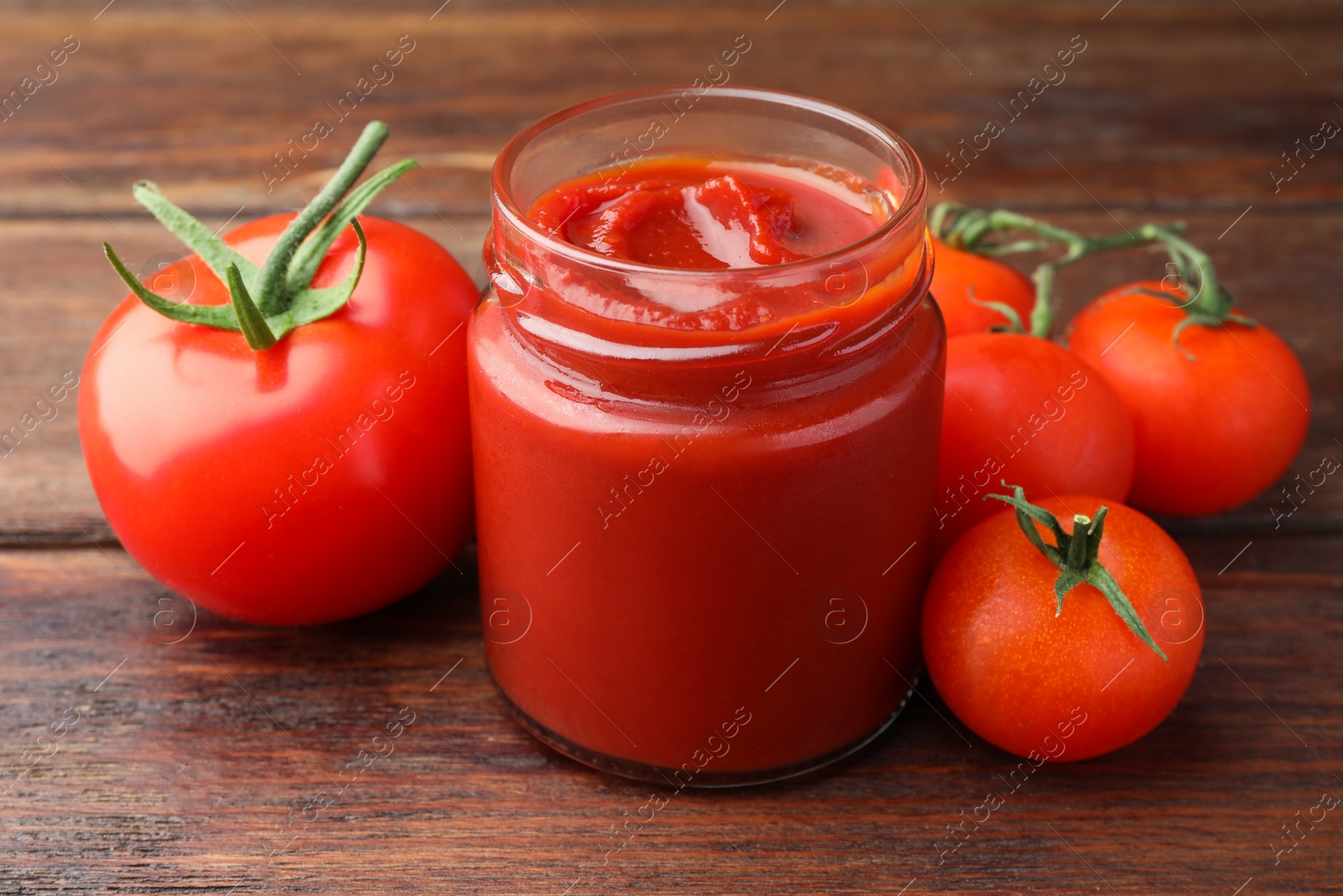 Photo of Jar of tasty ketchup and tomatoes on wooden table, closeup