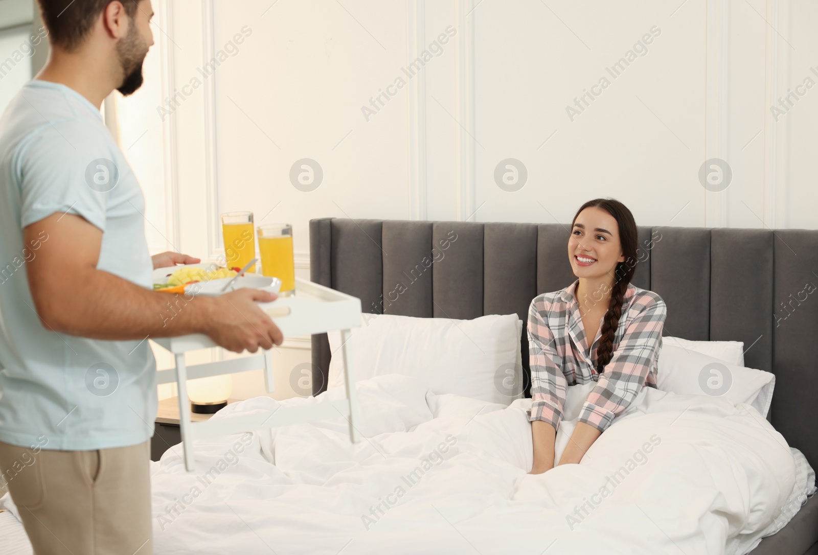 Photo of Man bringing white tray with breakfast to happy young woman on bed at home