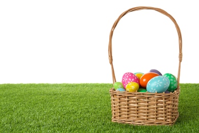 Photo of Wicker basket with Easter eggs on green grass against white background. Space for text