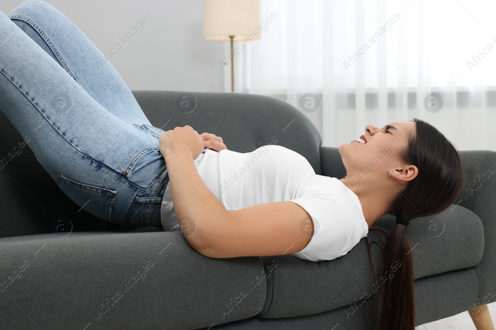 Photo of Young woman struggling to squeeze into tight jeans while lying on sofa at home