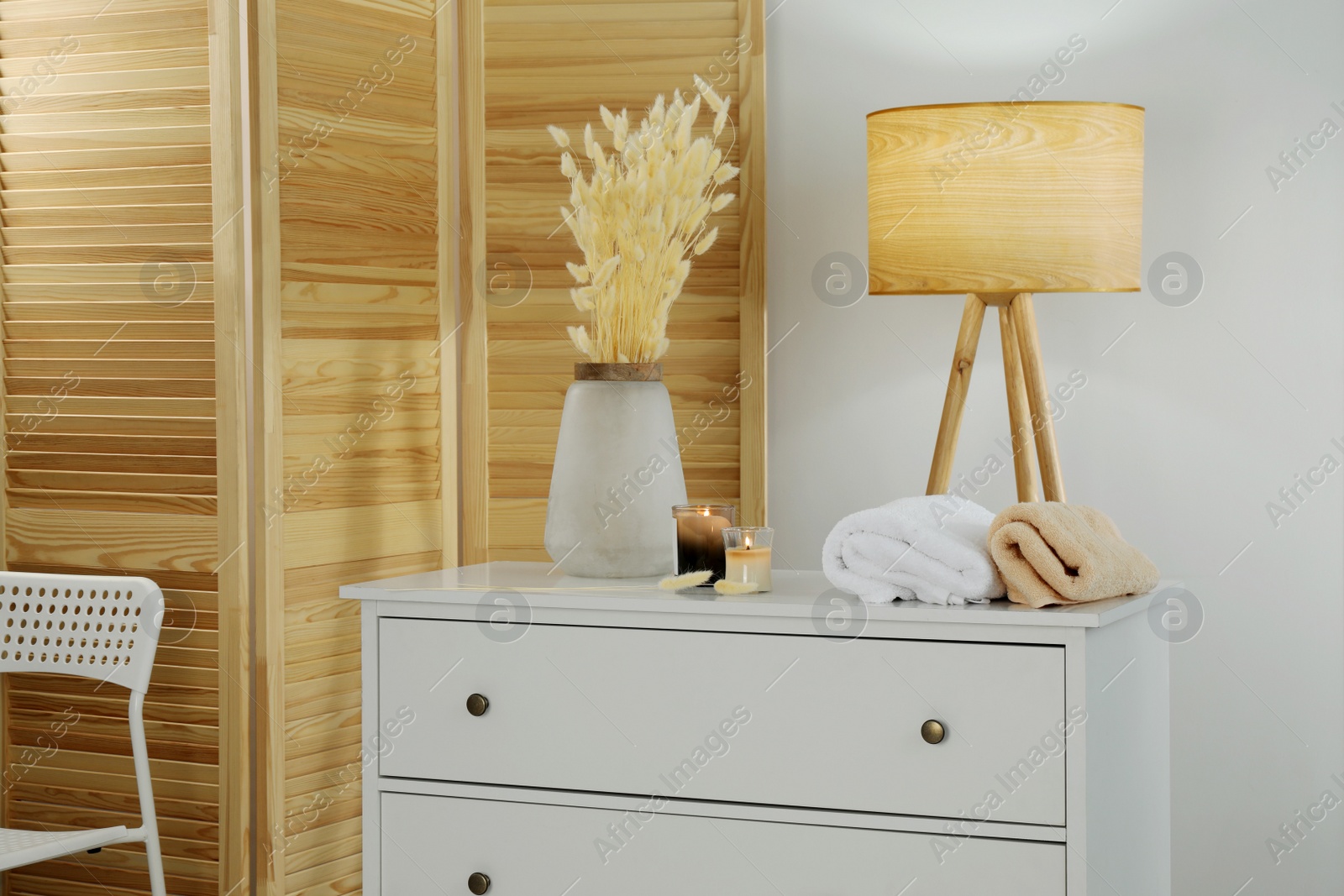 Photo of Vase with dried flowers, stacked towels and candles on chest of drawers in room. Interior design