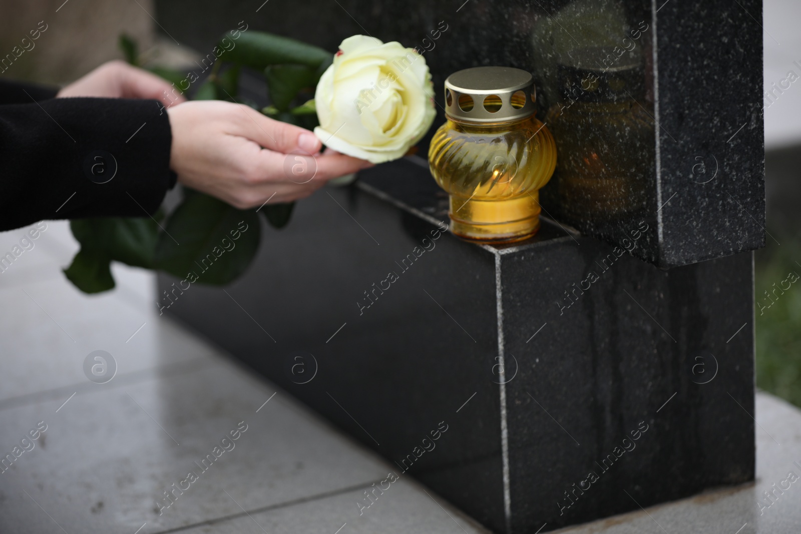 Photo of Woman holding white rose near black granite tombstone with candle outdoors, closeup. Funeral ceremony