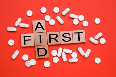Photo of Words First Aid made of wooden cubes and pills on coral background, flat lay
