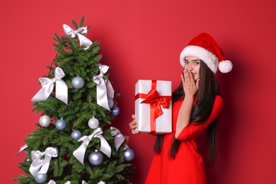 Beautiful young woman in Santa hat with gift box near Christmas tree on color background