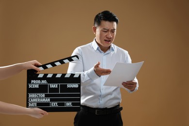 Photo of Emotional asian actor performing role while second assistant camera holding clapperboard on brown background. Film industry