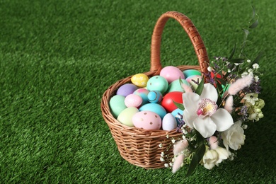 Photo of Wicker basket with Easter eggs and beautiful flowers on green grass. Space for text