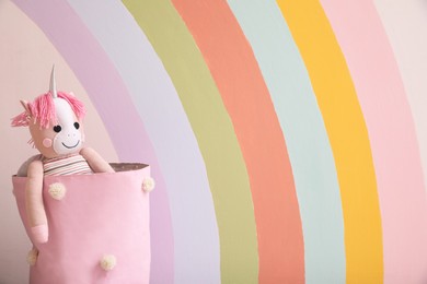 Toy unicorn in basket near wall with beautiful painted rainbow, space for text. Interior design