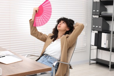 Photo of Young businesswoman waving hand fan to cool herself at table in office