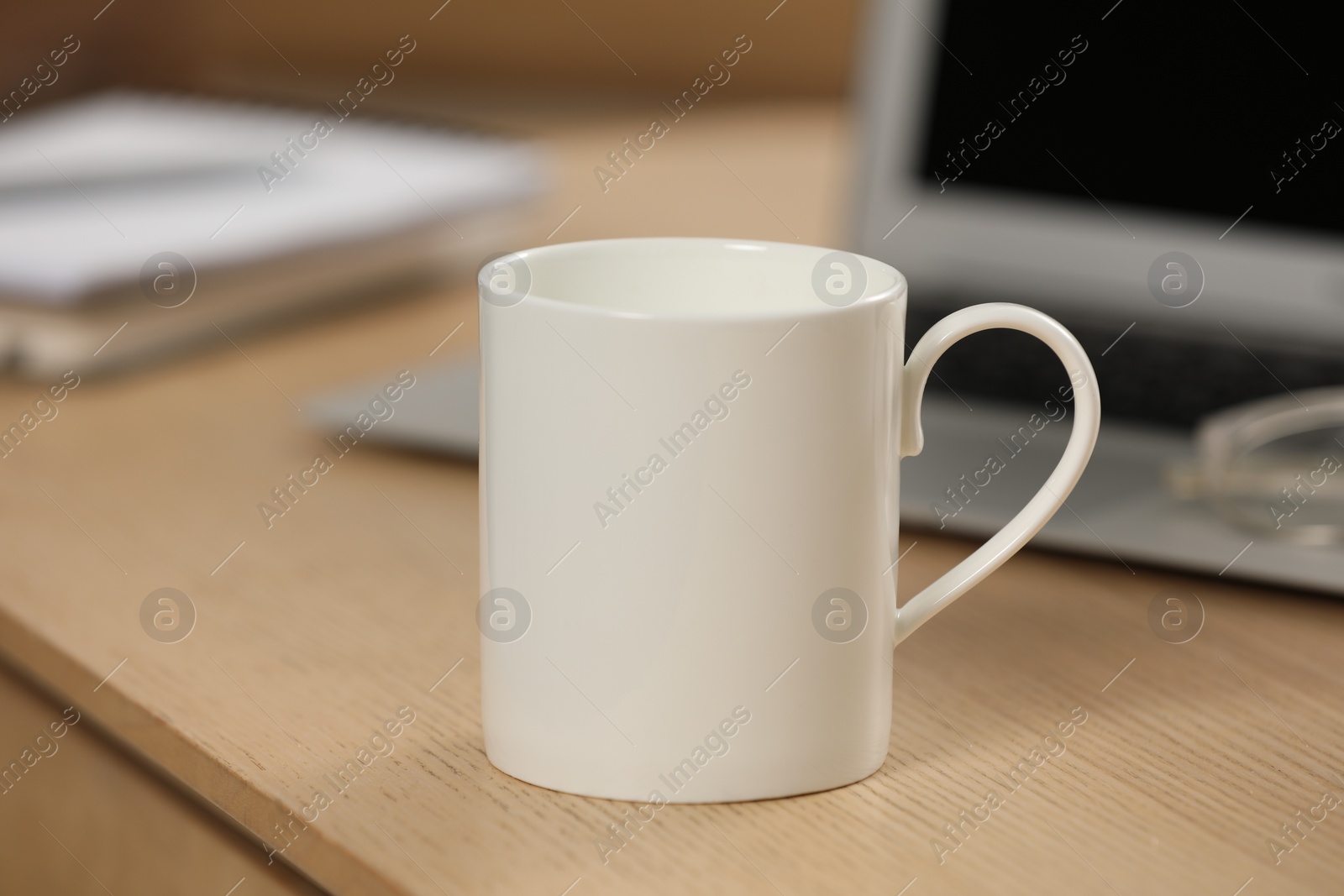 Photo of White ceramic mug and laptop on wooden table at workplace