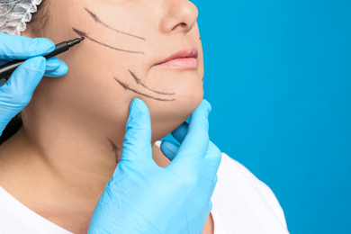 Doctor drawing marks on woman's face for cosmetic surgery operation against blue background, closeup. Double chin problem