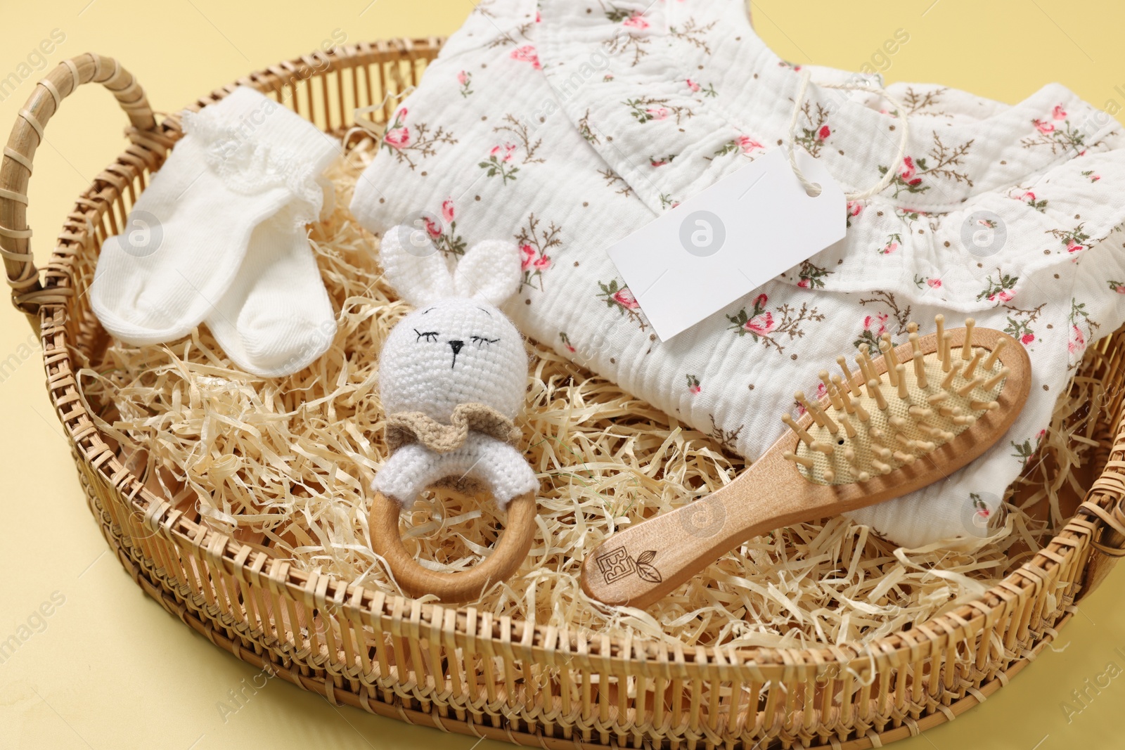 Photo of Different baby accessories, clothes and blank card in wicker basket on yellow background, closeup