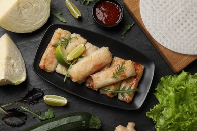 Photo of Tasty fried spring rolls, arugula, sauce and other products on dark textured table, flat lay