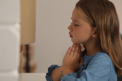 Photo of Cute little girl with hands clasped together praying at home. Space for text