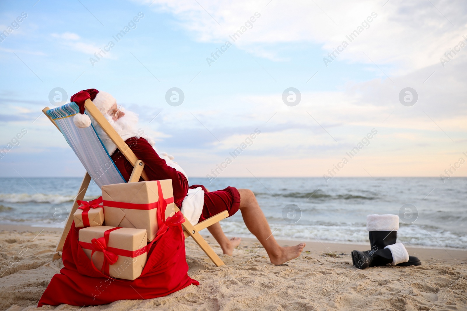 Photo of Santa Claus with bag of presents relaxing in chair on beach, space for text. Christmas vacation