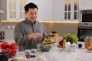 Photo of Cooking process. Man adding cut cucumber into bowl at countertop in kitchen