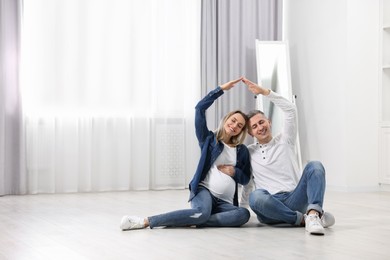 Pregnant woman with her husband forming roof with their hands while sitting on floor indoors. Space for text