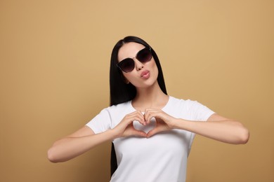 Photo of Beautiful young woman in stylish sunglasses blowing kiss and making heart with hands on beige background