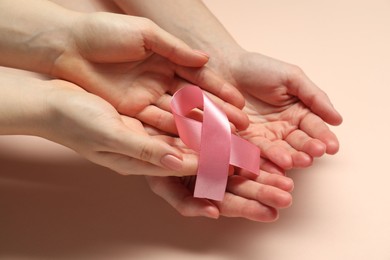 Women holding pink ribbon on beige background, closeup. Breast cancer awareness