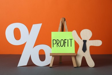 Photo of Economic profit. Wooden figure, easel with note and percent sign on grey table against orange background