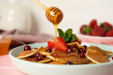Photo of Pouring honey onto cereal pancakes with berries on pink wooden table, closeup