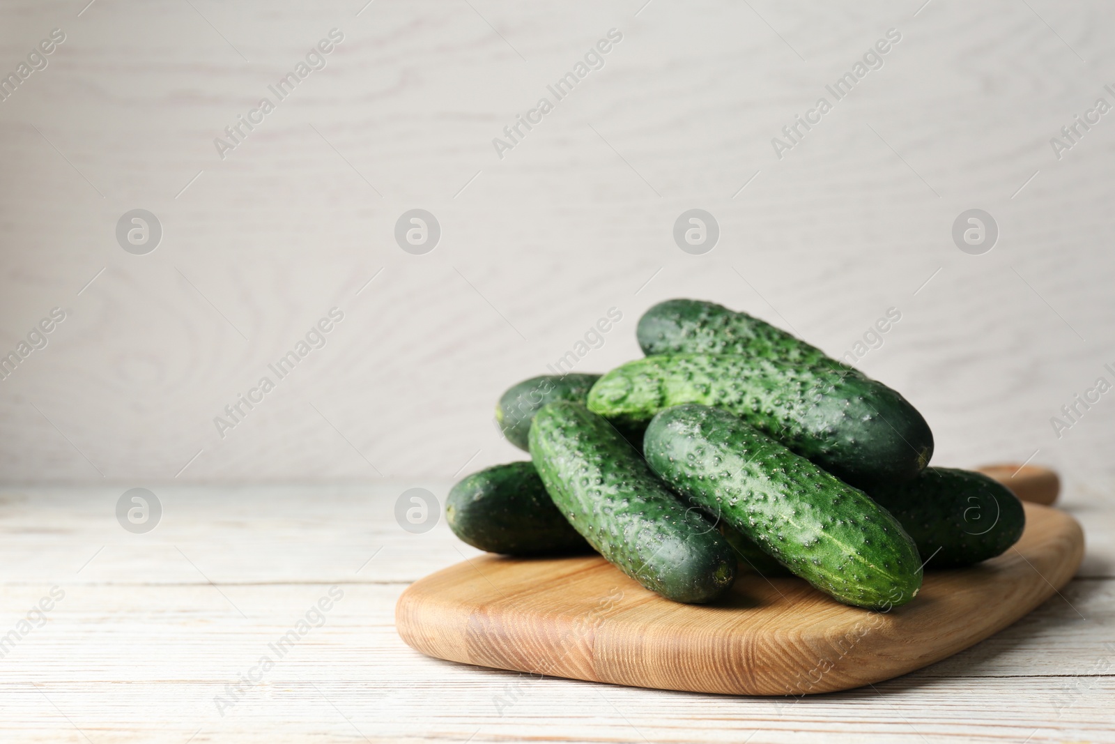 Photo of Board with fresh ripe cucumbers on white wooden table, space for text