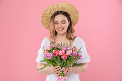 Happy young woman in straw hat holding bouquet of beautiful tulips on pink background