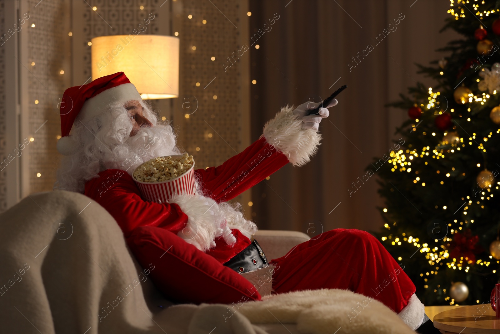 Photo of Merry Christmas. Santa Claus with popcorn bucket changing TV channels on sofa at home