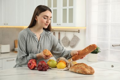 Photo of Woman with baguettes and string bag of fresh fruits at light marble table in kitchen
