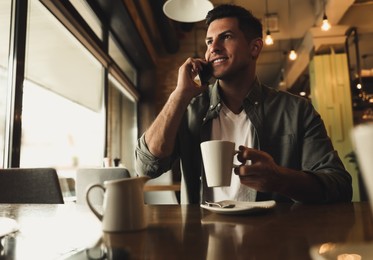 Photo of Handsome man with cup of coffee talking on smartphone at cafe in morning