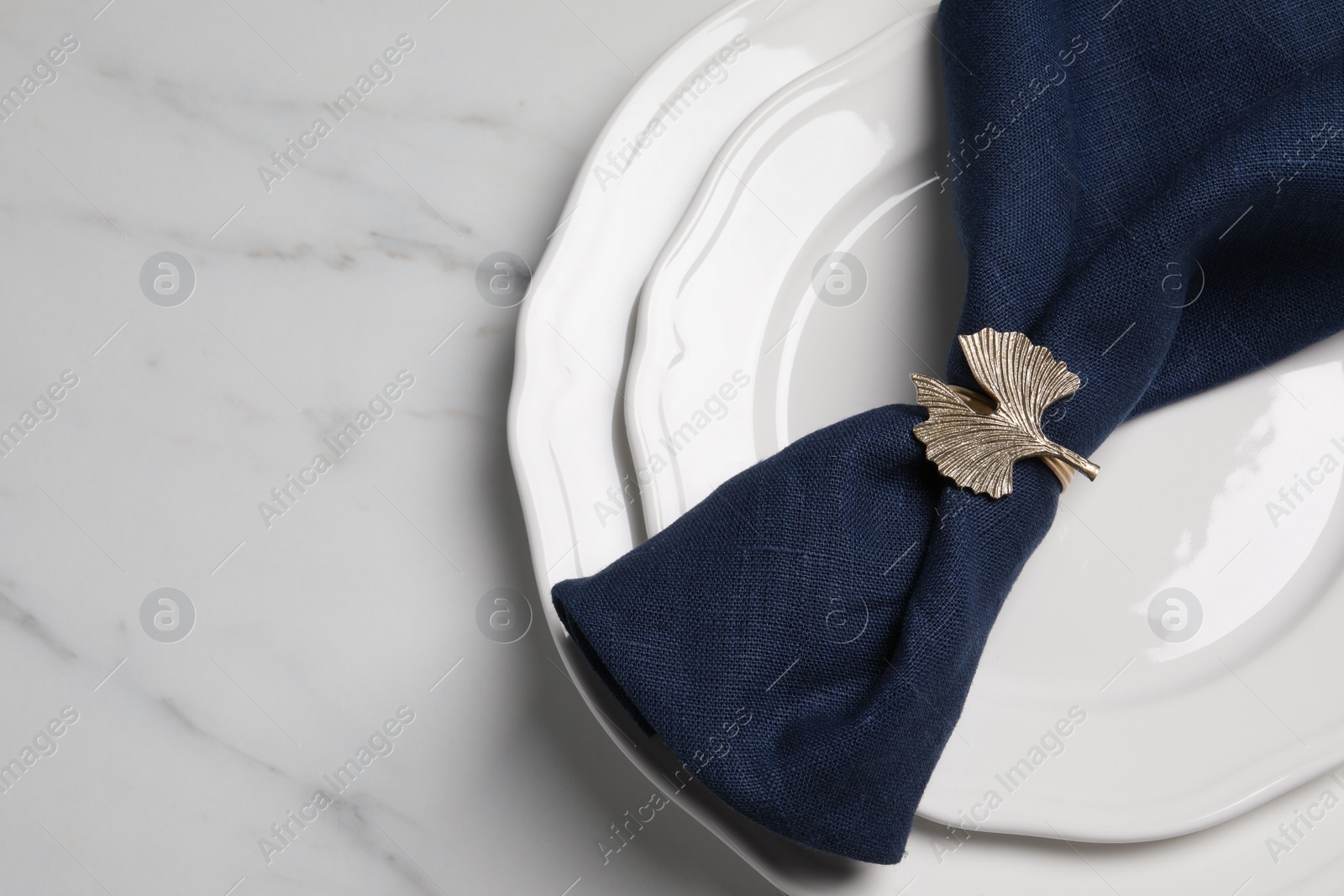 Photo of Plate with dark blue fabric napkin and decorative ring on white marble table, top view. Space for text