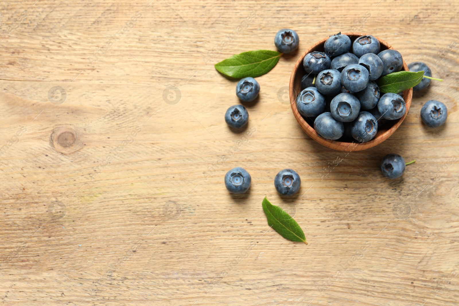 Photo of Bowl of tasty fresh blueberries with leaves on wooden table, flat lay. Space for text