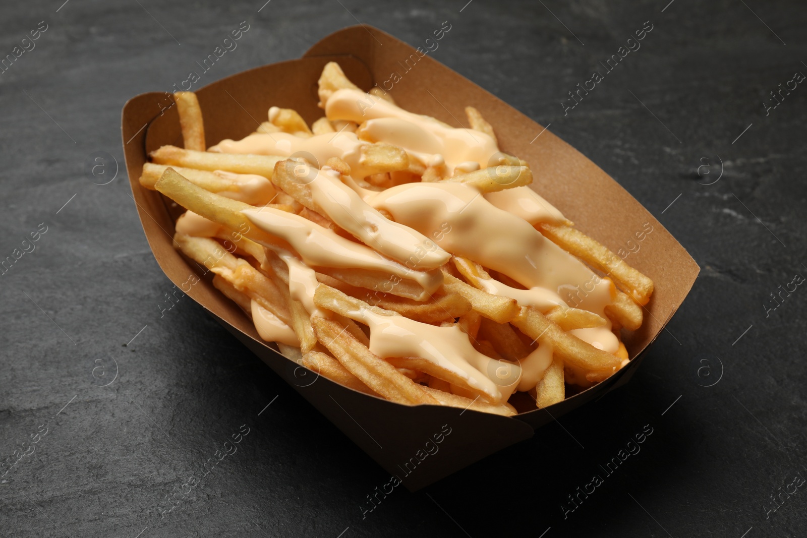 Photo of Tasty potato fries and cheese sauce in paper container on black table, closeup