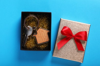 Key with trinket in shape of house and gift box on light blue background, flat lay. Housewarming party