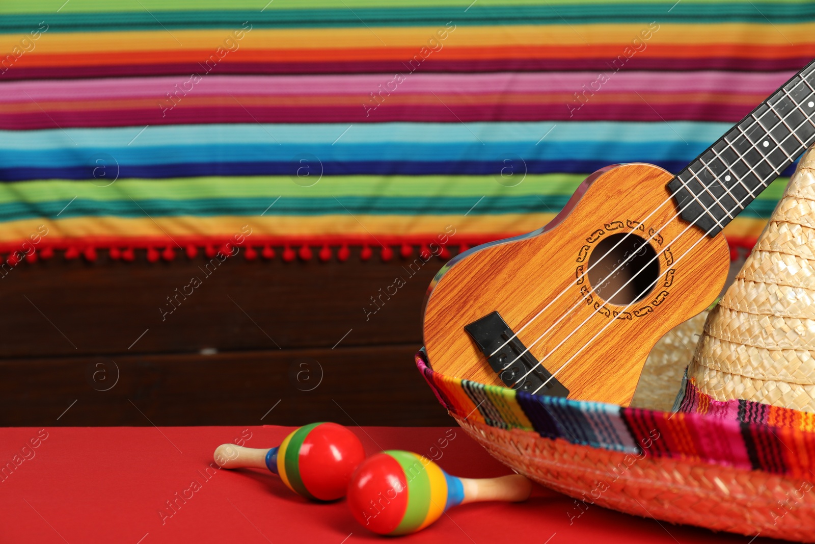 Photo of Mexican sombrero hat, ukulele and maracas on red table, closeup. Space for text