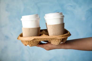 Woman holding cardboard holder with takeaway paper coffee cups on light blue background, closeup