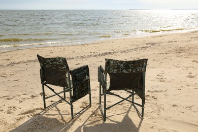 Comfortable camouflage fishing chairs on sandy beach near sea