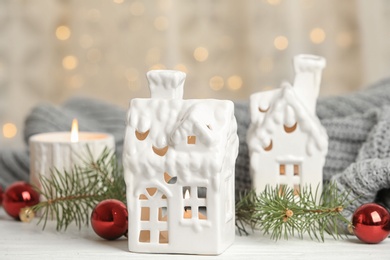 Photo of House shaped holders with burning candles on white wooden table against blurred Christmas lights